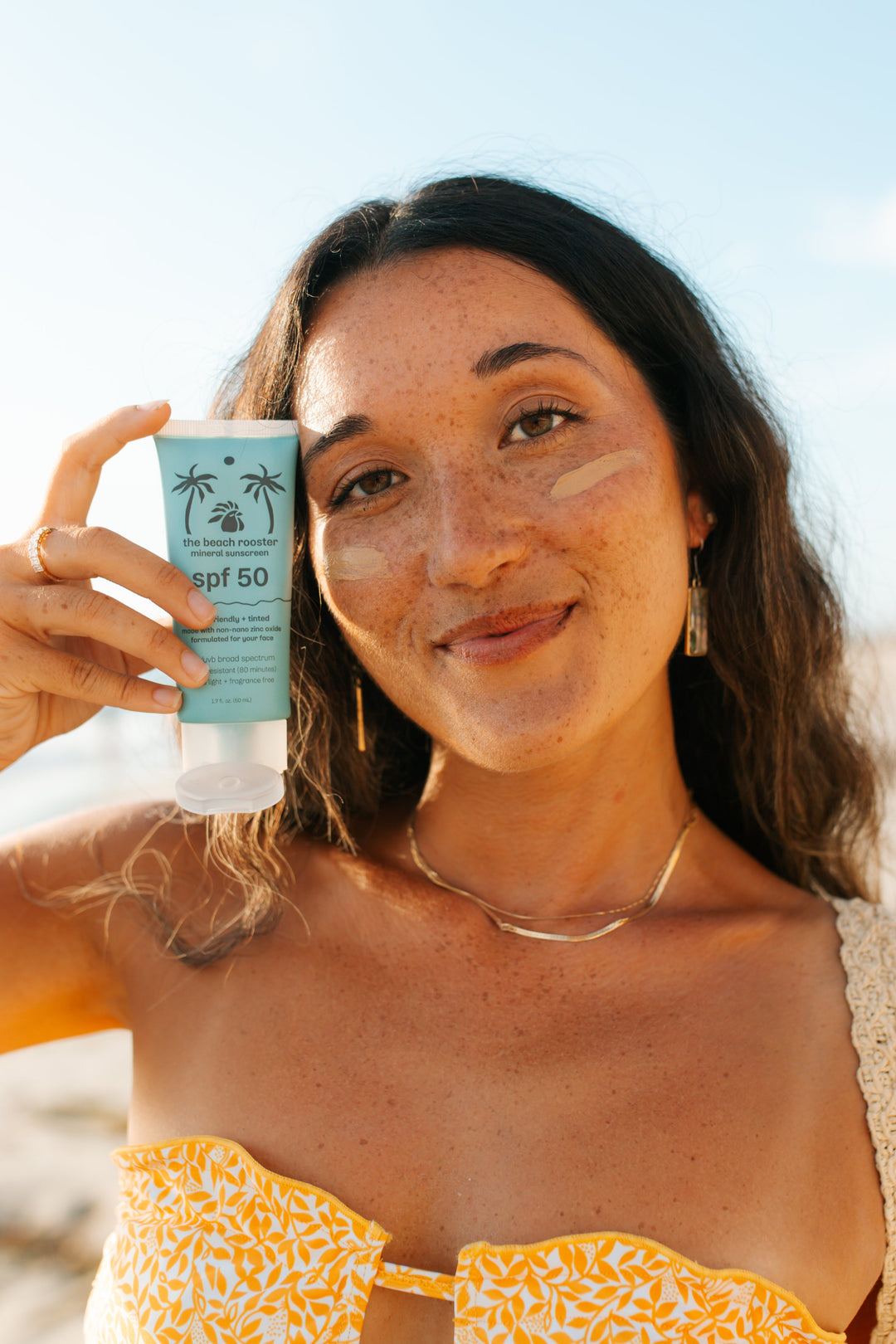A woman on a beach holds up a tube of tinted mineral sunscreen next to her face, which has SPF 50 coverage. The sunscreen tube is light blue with a black the beach rooster.