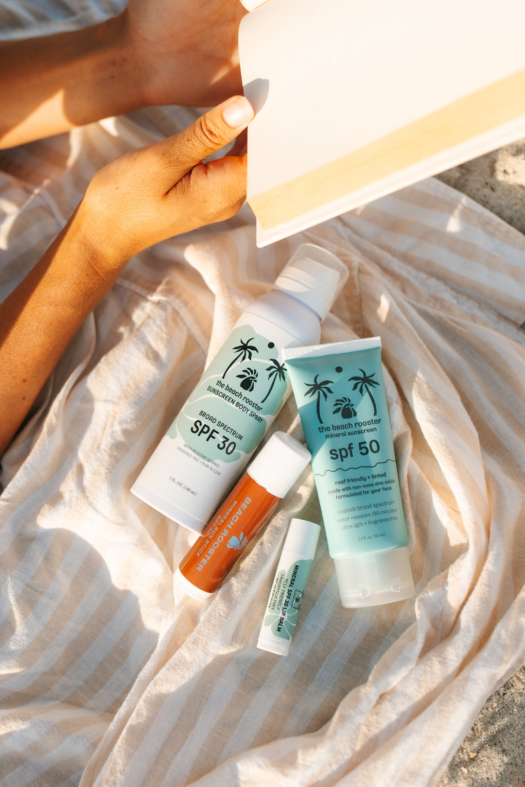 Sun protection products from The Beach Rooster are arranged on a striped beach towel outdoors, with a person's hand holding a book nearby. 