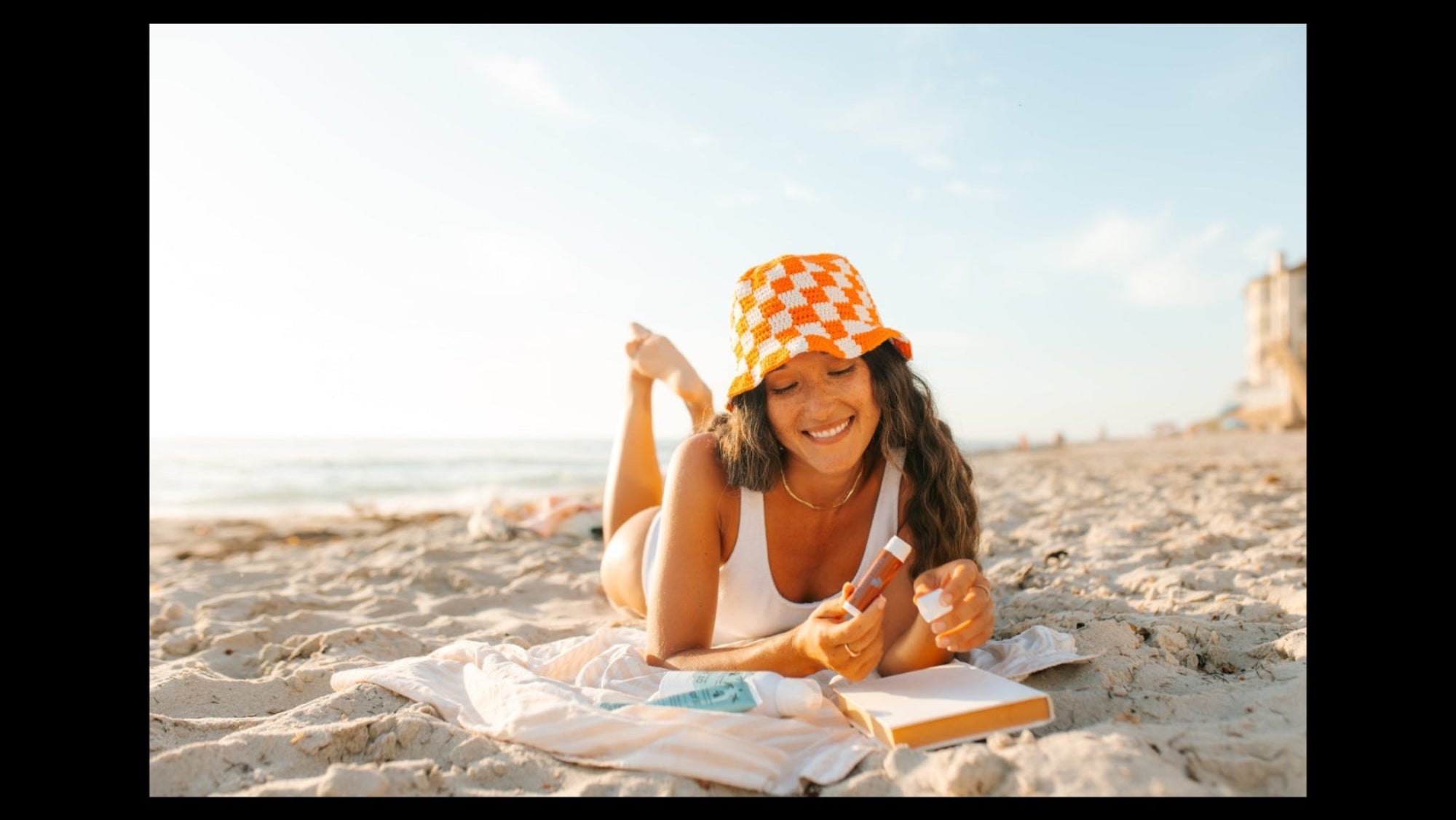 A woman lies on a sandy beach wearing a white bathing suit and an orange-and-white checkered bucket hat, holding sunscreen and an open notebook under a bright sky.