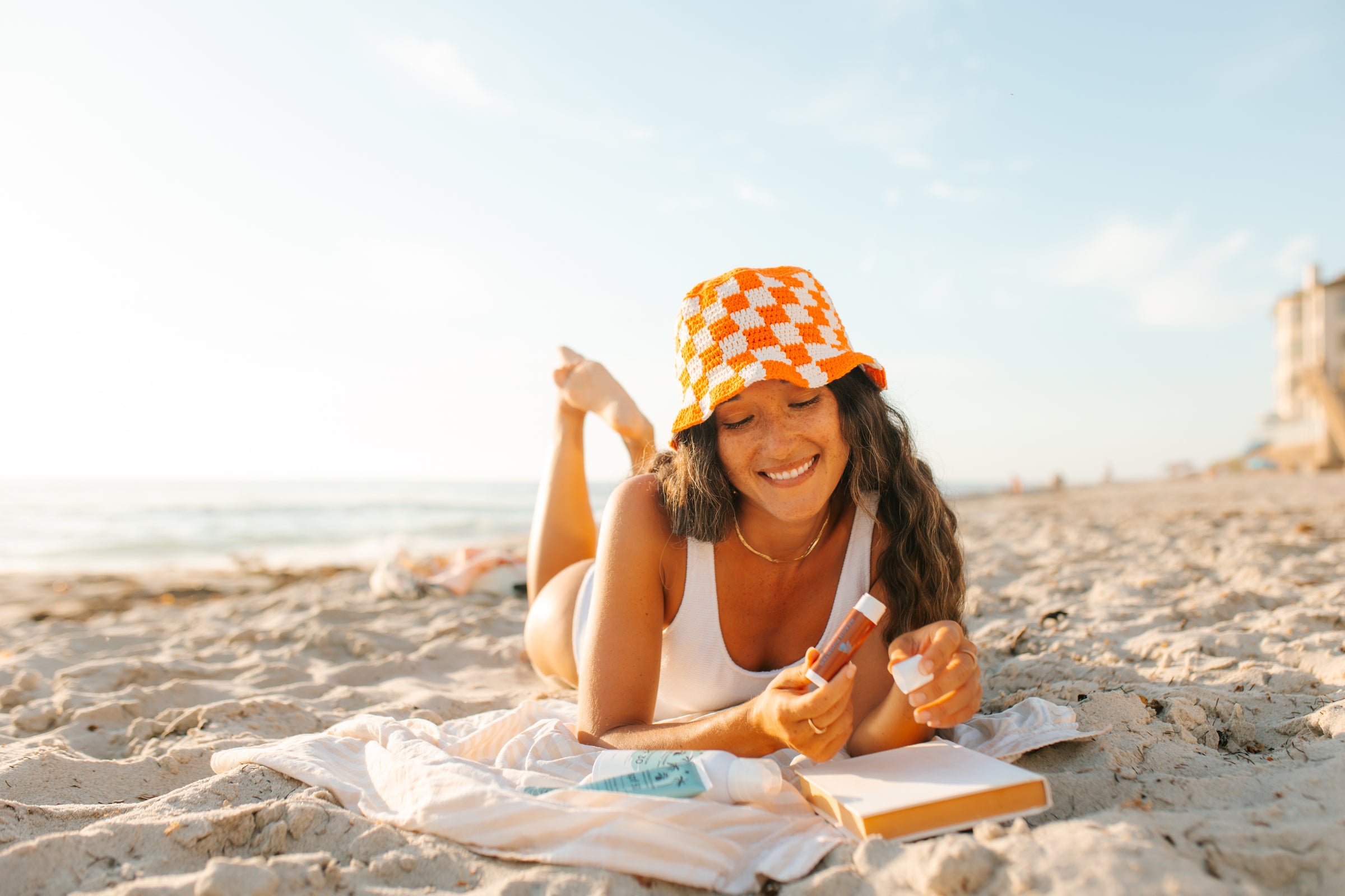 A woman lies on a sandy beach wearing a white bathing suit and an orange-and-white checkered bucket hat, holding sunscreen and a notebook under a bright sky.