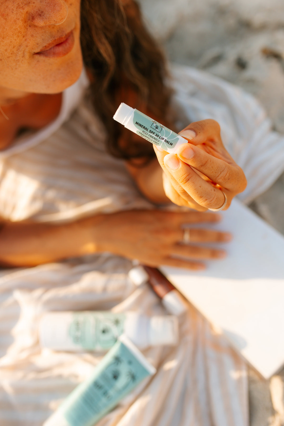A close-up lifestyle shot of a woman holding a small sunscreen lip balm on a beach. The tube is light blue and white with the brand name the beach rooster.