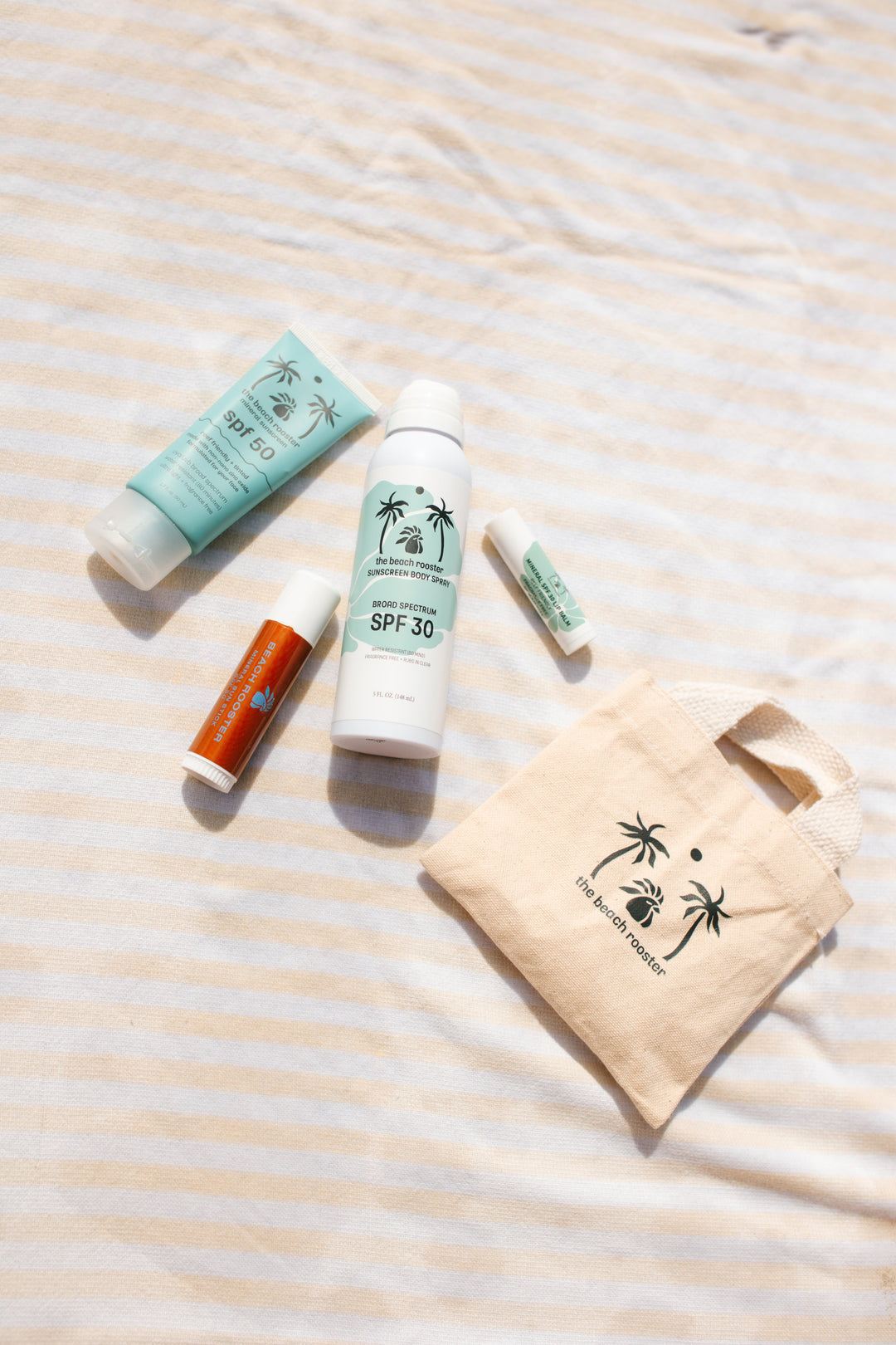 A sunscreen and lip care set from The Beach Rooster, including a light blue tube of mineral sunscreen SPF 50, a white bottle of broad spectrum sunscreen body spray SPF 30, and two lip products on a striped towel with a small tote bag with the beach rooster logo on it. 