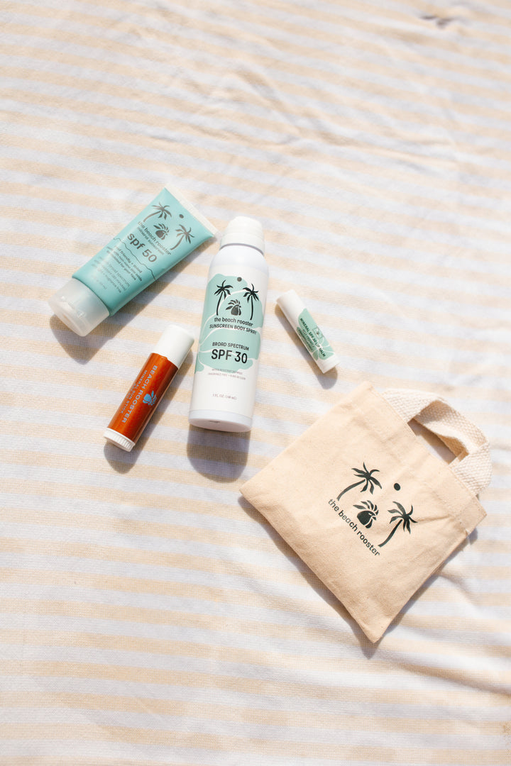 A sunscreen and lip care set from The Beach Rooster, including a light blue tube of mineral sunscreen SPF 50, a white bottle of broad spectrum sunscreen body spray SPF 30, and two lip products on a striped towel with a small tote bag with the beach rooster logo on it. 