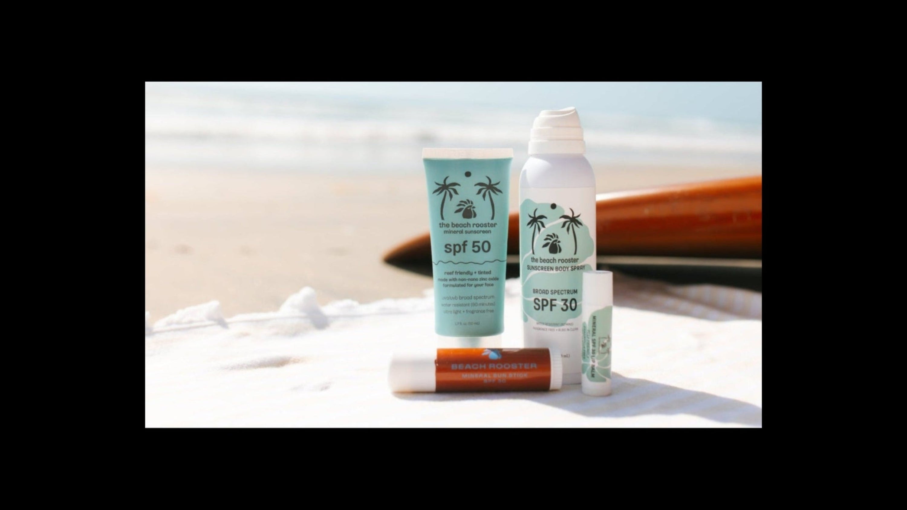 Four sunscreen and lip care products from The Beach Rooster are arranged on a white towel on a sandy beach with the ocean and sky blurred in the background. The products include SPF 50, SPF 30, and a lip balm.