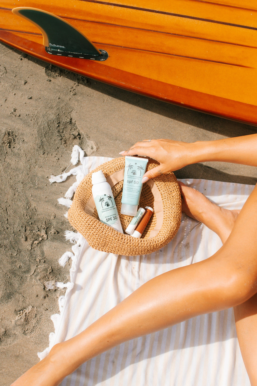 A sunscreen and lip care set from The Beach Rooster, including a light blue tube of mineral sunscreen SPF 50, a white bottle of broad spectrum sunscreen body spray SPF 30, and two lip products, arranged in a straw hat sitting on a white towel with a surfboard in the background.