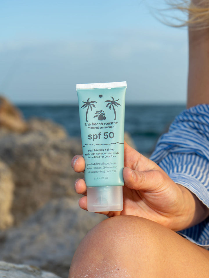 Woman holding The Beach Rooster SPF 50 mineral suncreen with a rocky beach and sky background 