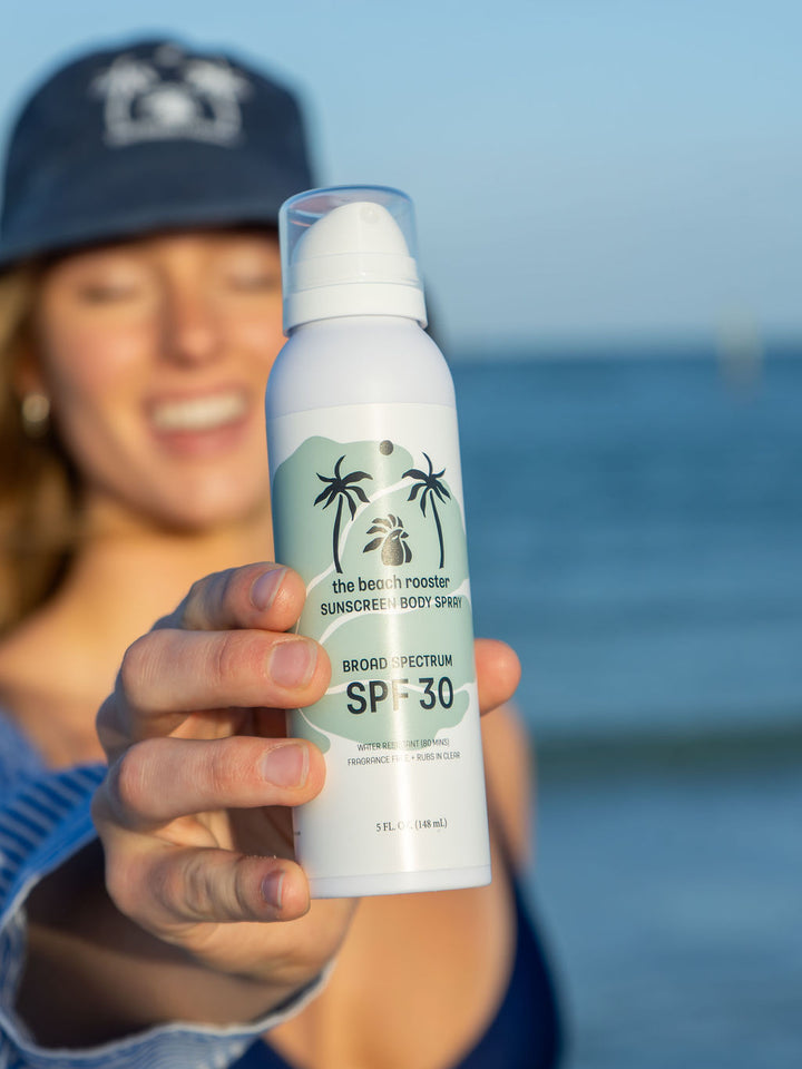 Woman holding The Beach Rooster SPF 30 Mineral Sunscreen Body Spray on a beach 