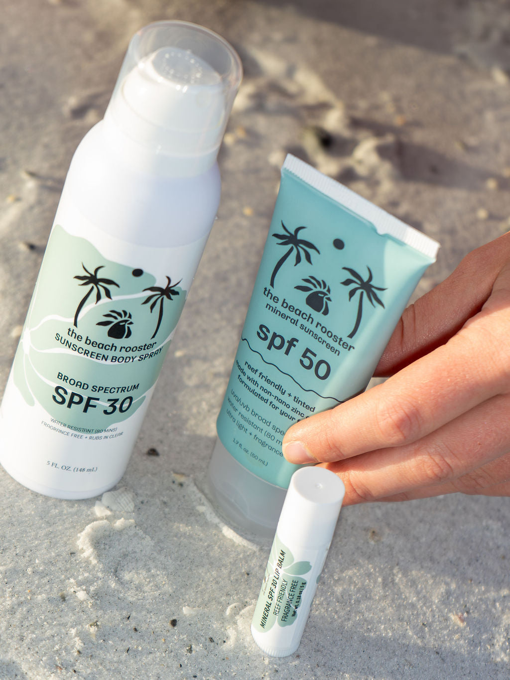 The Beach Rooster Bundle SPF Mineral Sunscreen, Face, Body and Lips with a sandy and beach background and a woman hand reaching for the face spf 
