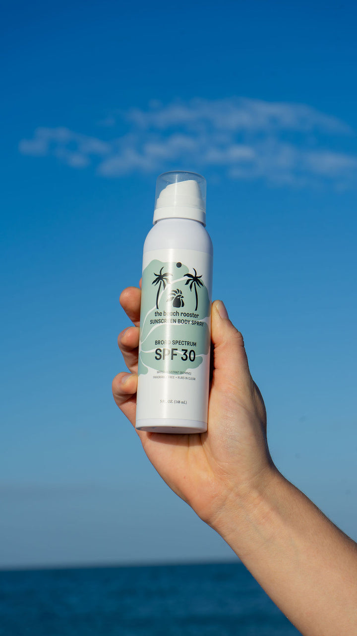 Woman holding The Beach Rooster SPF 30 Mineral Sunscreen Body Spray with a blue sky background 