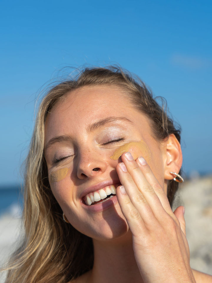 Smiling woman putting on The Beach Rooster SPF 50 mineral suncreen to her face 