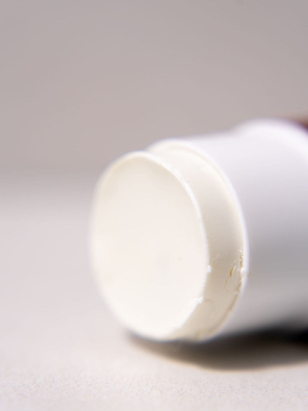 A close-up, shallow depth-of-field shot of a white cosmetic stick, showing the rounded end cap with a slightly textured surface, resting on a neutral light gray background.