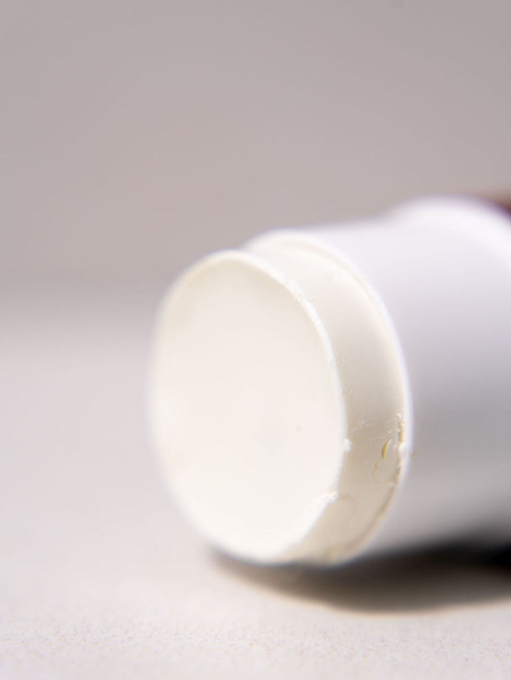 A close-up, shallow depth-of-field shot of a white cosmetic stick, showing the rounded end cap with a slightly textured surface, resting on a neutral light gray background.