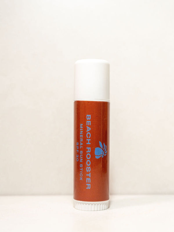 A single tube of Beach Rooster Mineral Sun Stick lip balm, featuring a terracotta orange-brown body with blue branding and a white cap, standing upright on a white surface against a white background.