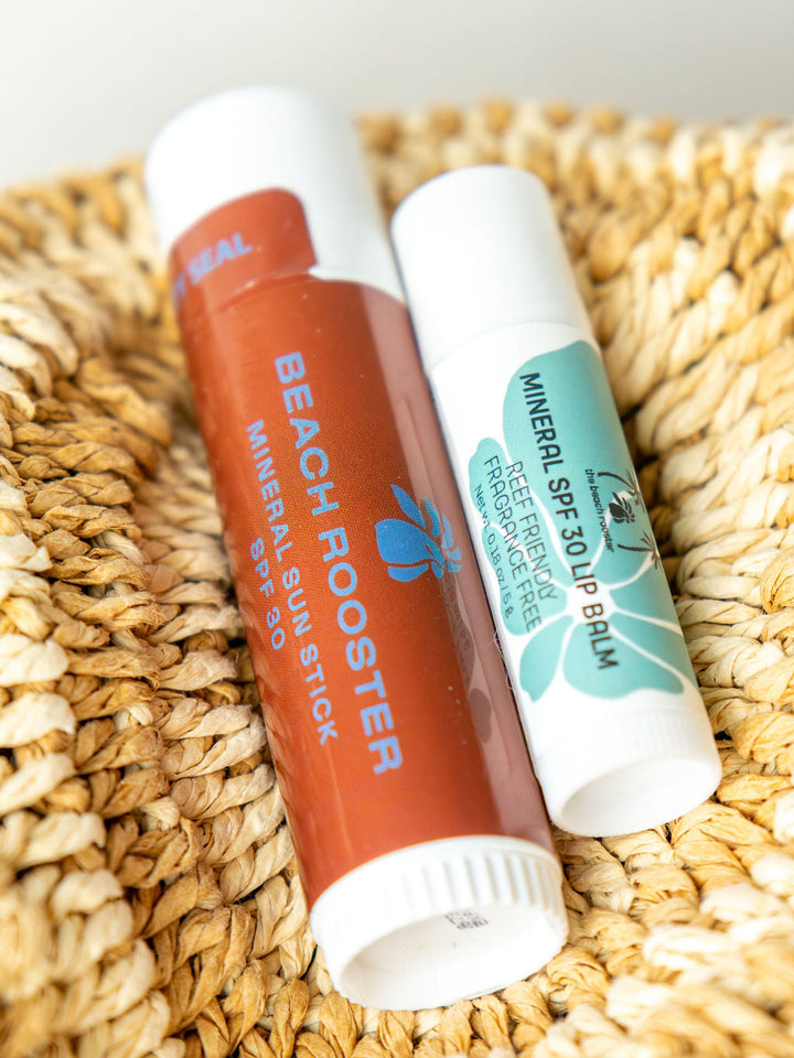 One lip balm and one face sunscrren stick resting on a woven straw mat. 