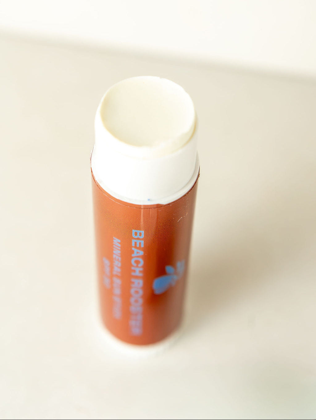 A single tube of face sunscreen standing upright on a light surface against a white background. The tube is a warm terracotta orange-brown color with a white cap, and the text reads 'BEACH ROOSTER' with a small blue bird graphic.

