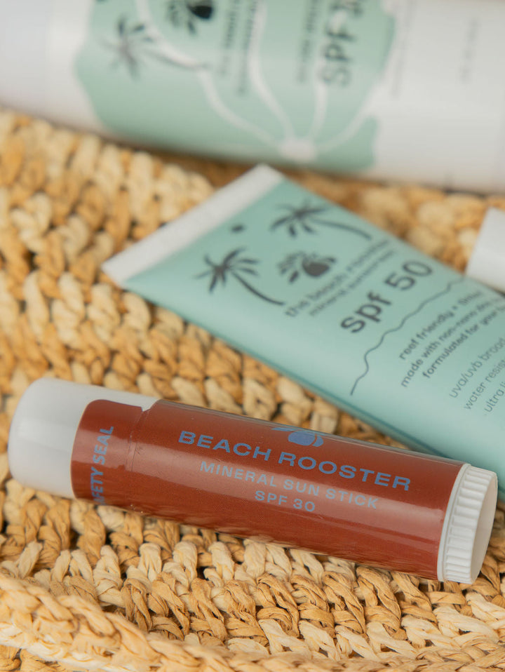 A face sunscreen tube 'BEACH ROOSTER' in the foreground on a woven straw mat, with SPF 50 sunscreen tubes behind it in a styled product shot.

