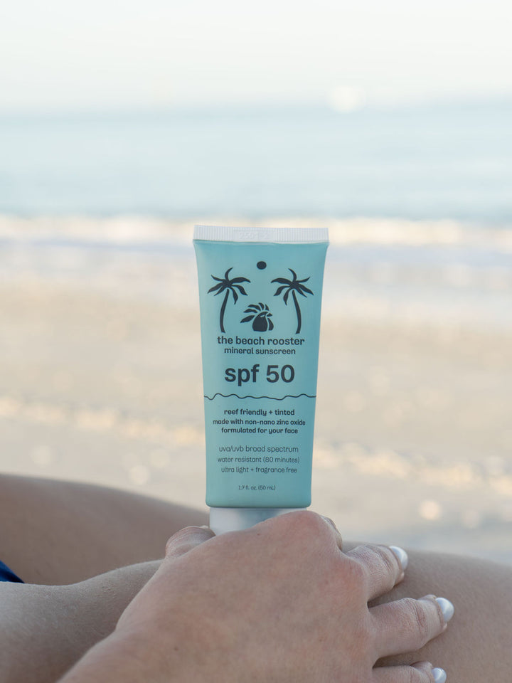 Woman holding The Beach Rooster SPF 50 mineral suncreen with a beach and sky background 