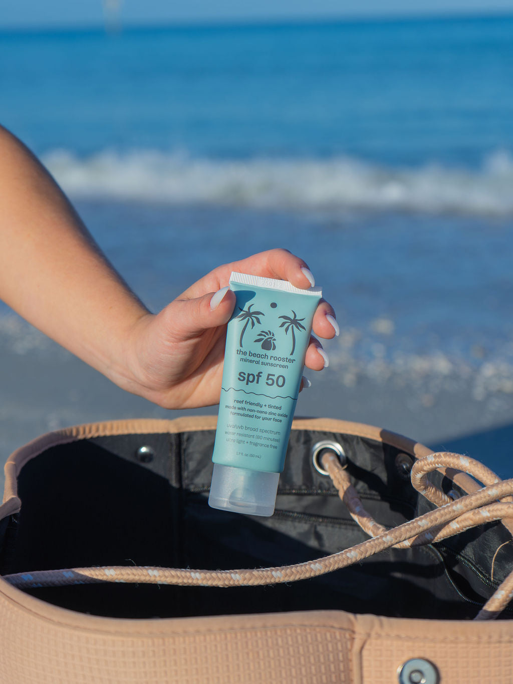 Woman holding The Beach Rooster SPF 50 mineral suncreen and packing it into her beach bag at a beach with water and sky in the background 