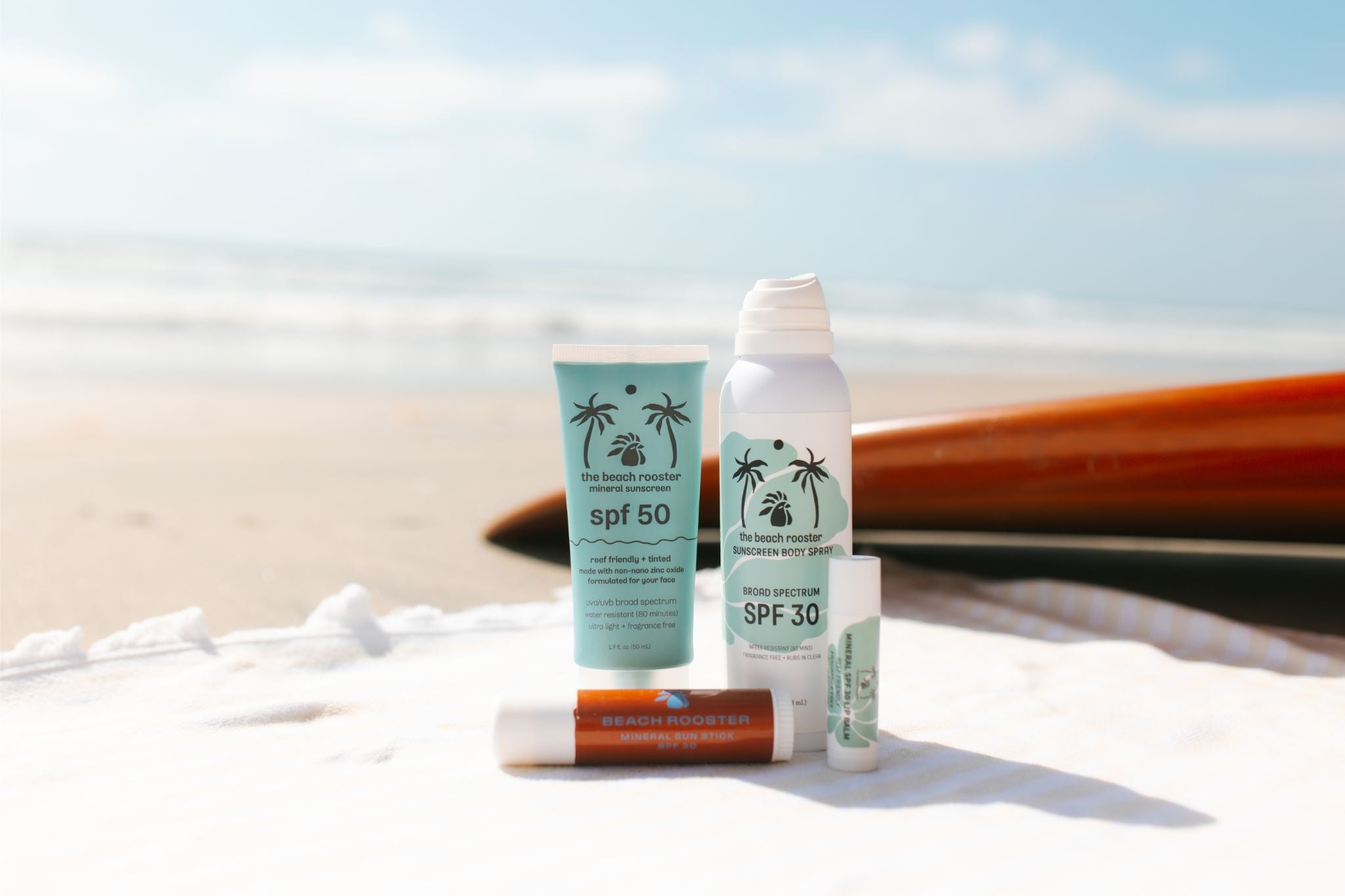 A set of sunscreen and lip care products from The Beachy Rooster Company arranged on a white towel at the beach, with the ocean and sand blurred in the background and a wooden surfboard behind them. The products include an SPF 50 tube, an SPF 30 spray bottle, a lip balm tube, and a face stick .