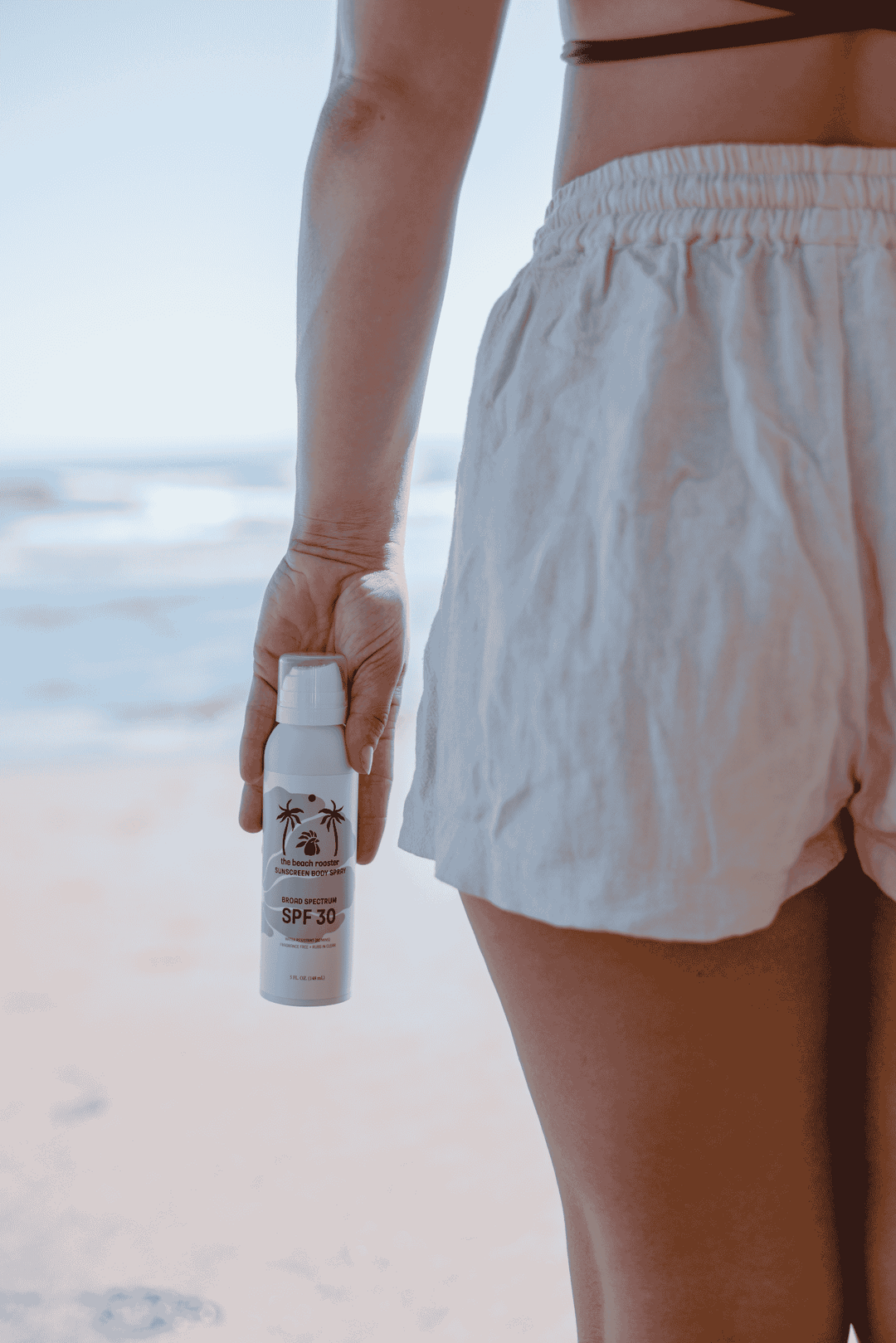 Woman holding a mineral sunscreen spray with a beach and ocean background
